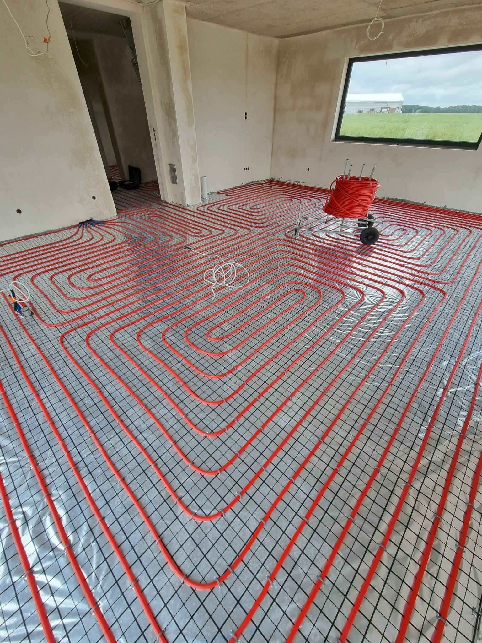 Czerwone rury ogrzewania podłogowego ułożone na srebrnej folii z czarną siatką w surowym wnętrzu z oknem i rolką rur na wózku. Widok z góry.