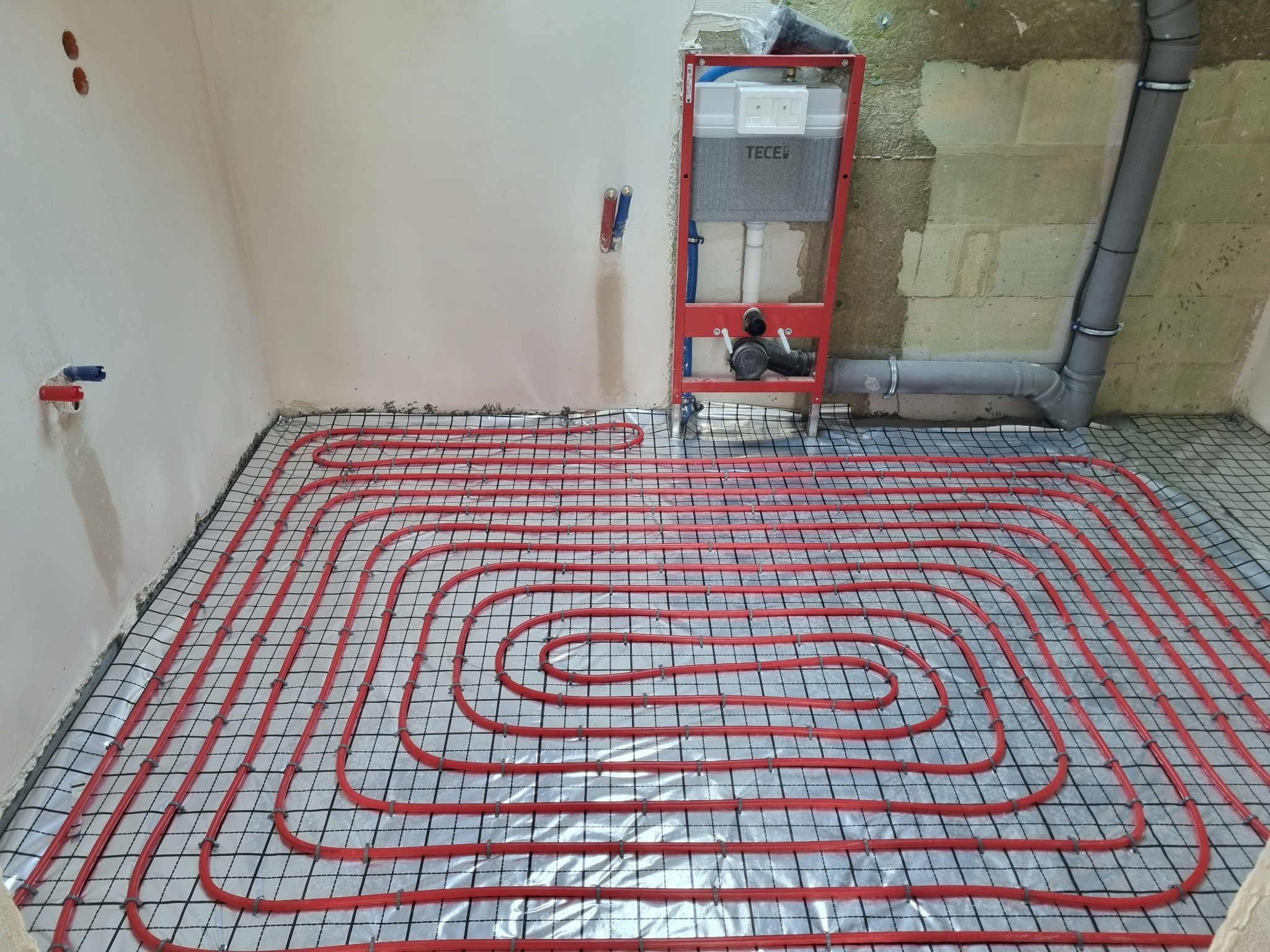 Czerwone rury ogrzewania podłogowego ułożone na folii z rastrem, widoczny stelaż podtynkowy WC TECE i szare rury kanalizacyjne. Etap prac instalacyjnych.