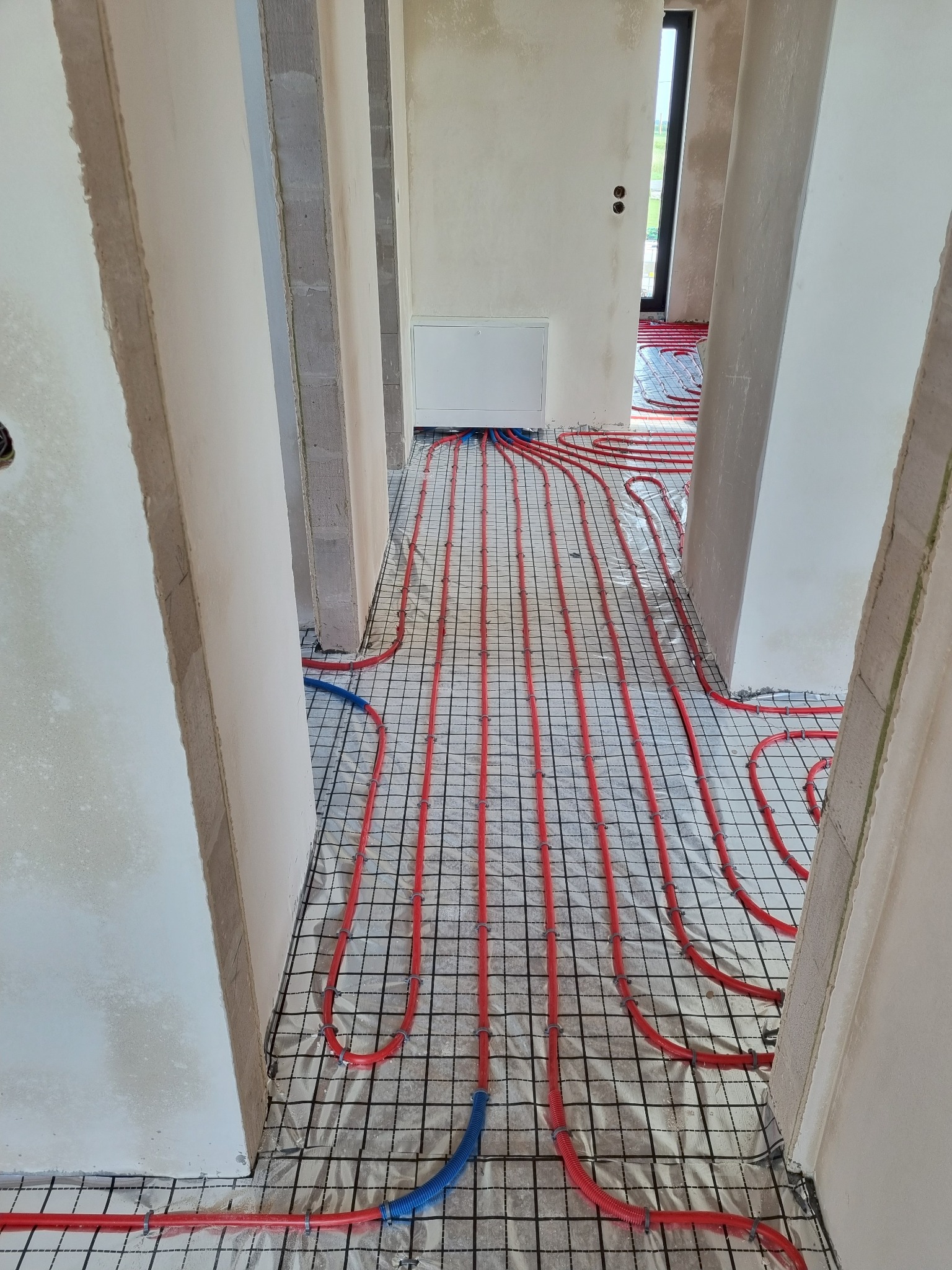 Ułożone czerwone rury ogrzewania podłogowego na folii z siatką w surowym wnętrzu. Widoczne nieotynkowane ściany i fragment okna. Etap prac instalacyjnych.