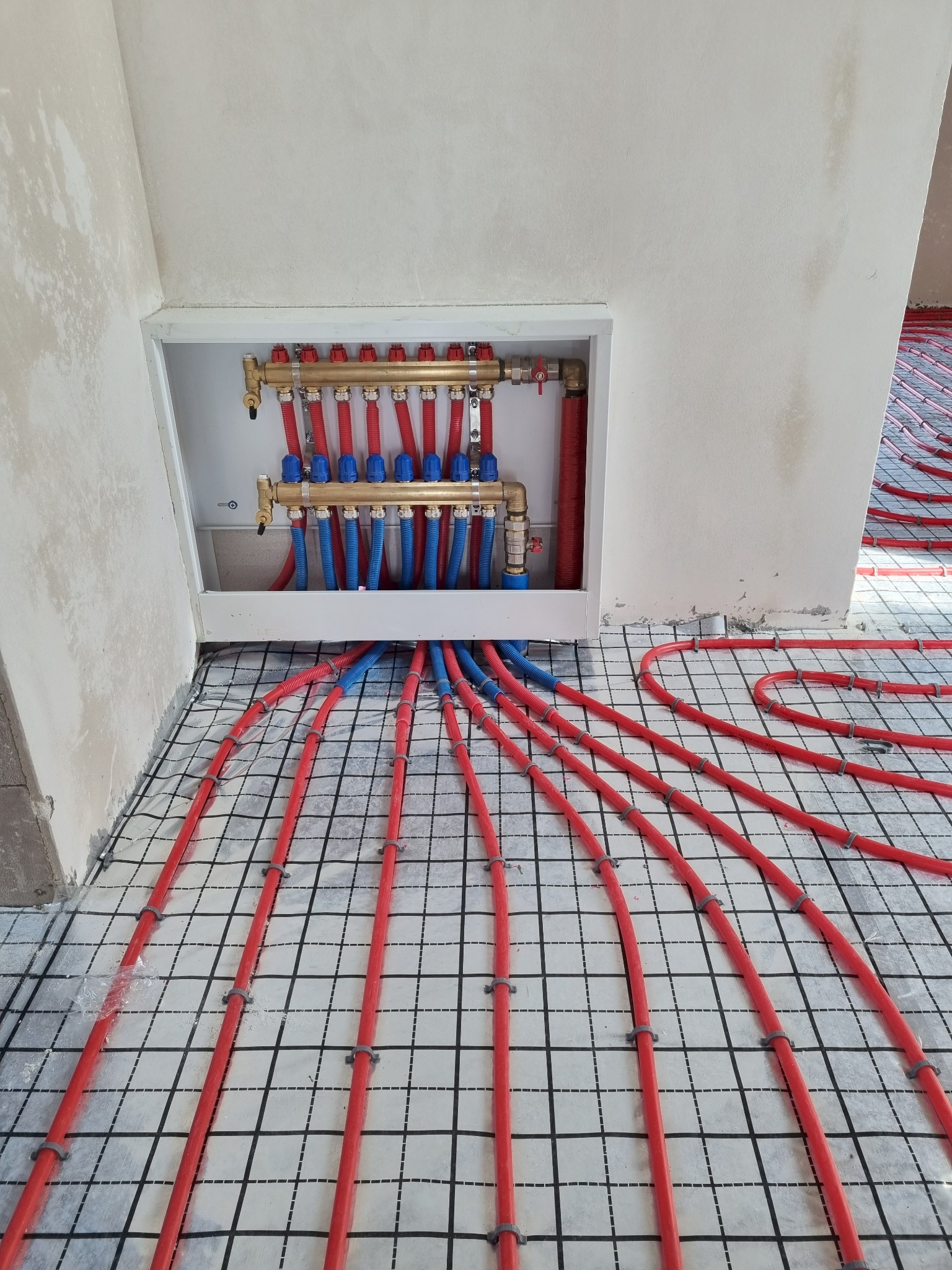Rozłożona instalacja ogrzewania podłogowego z czerwonymi rurami na folii z rastrem, widoczny rozdzielacz wnękowy z niebieskimi i czerwonymi elementami. Świeżo położona instalacja.