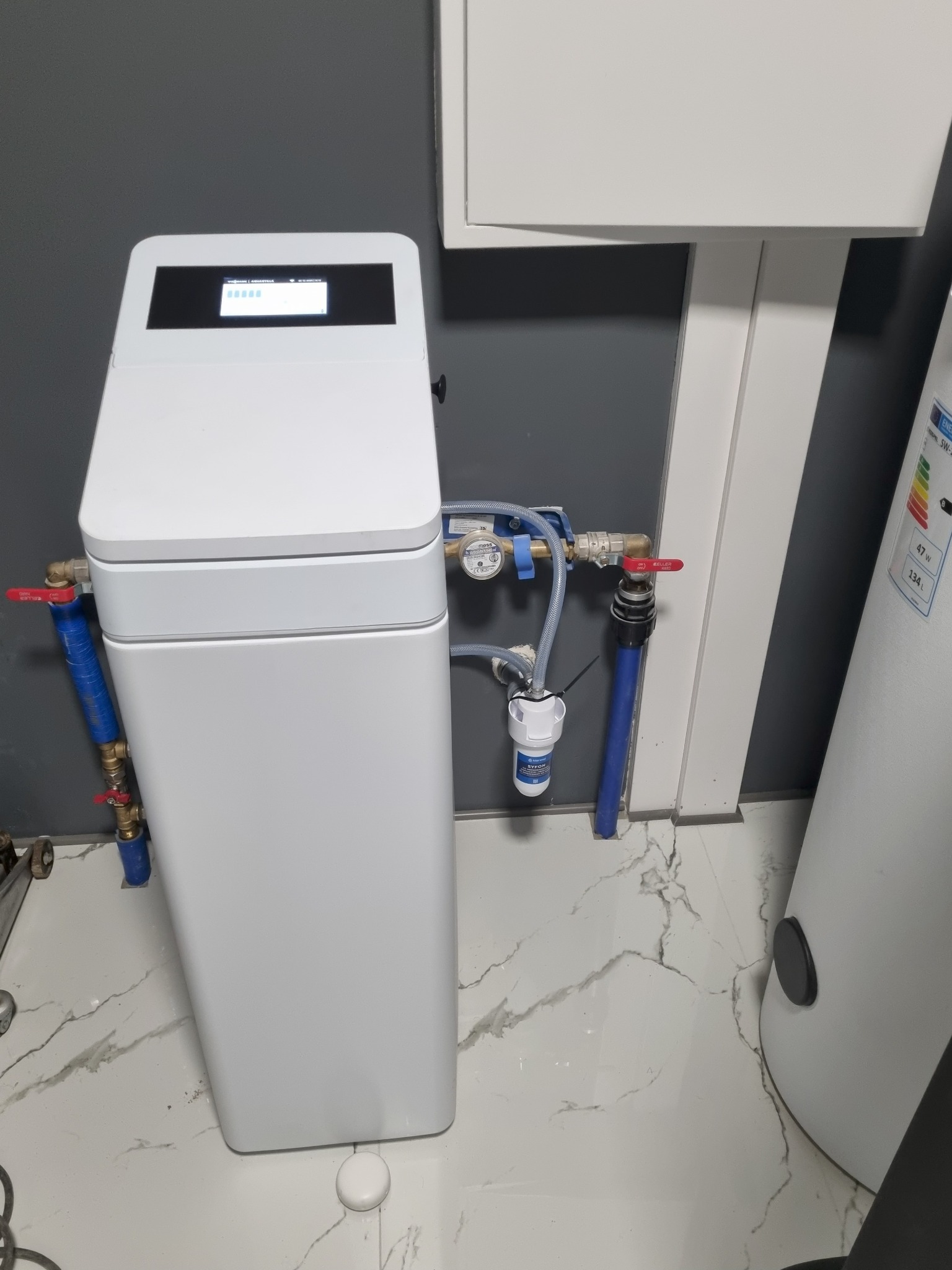 Biały zmiękczacz wody z podłączonymi rurami i manometrem, zainstalowany przy szarej ścianie i białym bojlerze na marmurowej podłodze. Widoczne zawory i filtr.