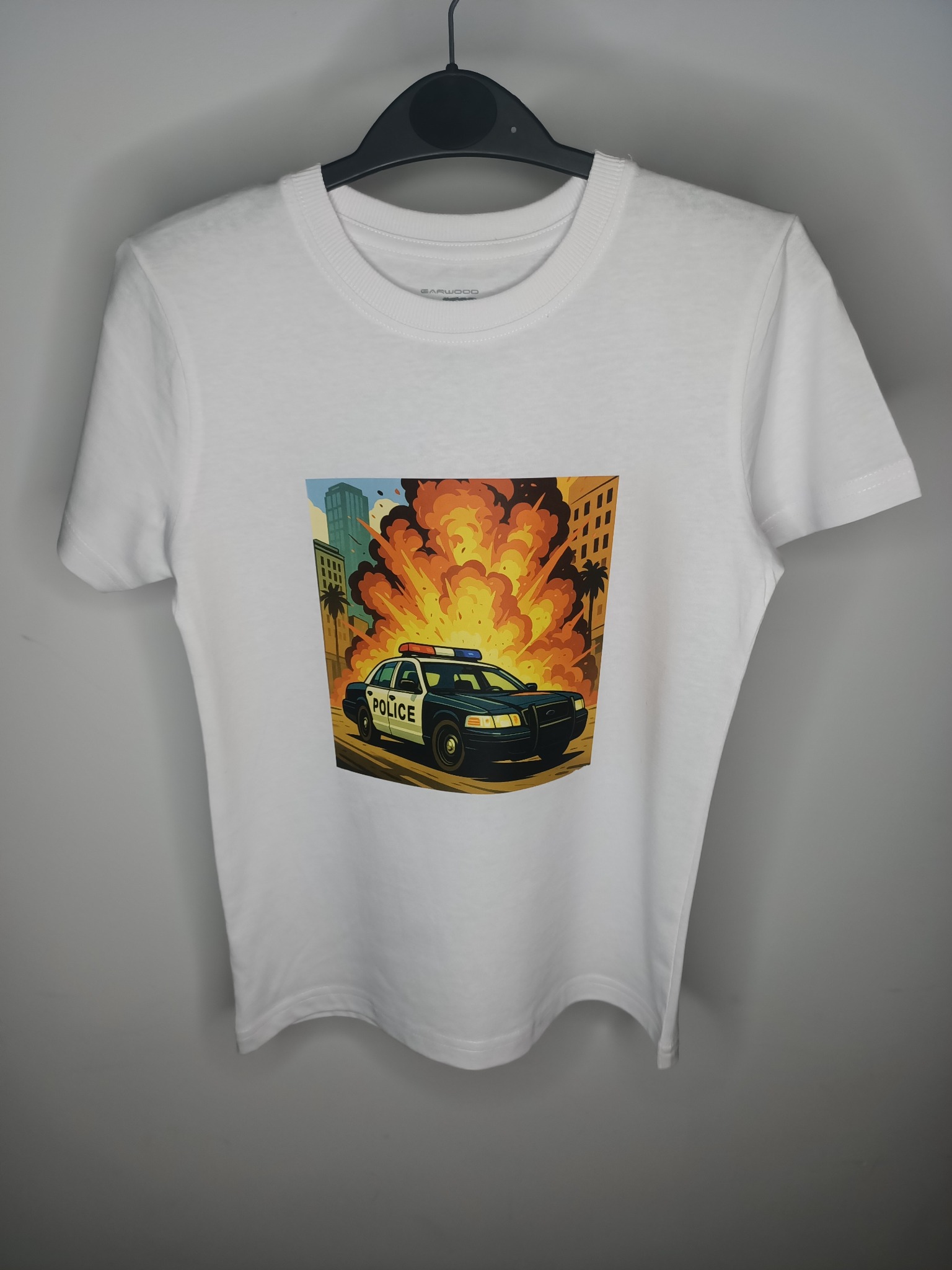 Biała koszulka z krótkim rękawem, na wieszaku, z nadrukiem przedstawiającym radiowóz policyjny na tle eksplozji w mieście. Grafika w stylu komiksowym.