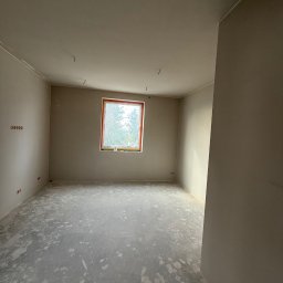 REMIGIUSZ PUKACKI REMIPOL - Wnętrze pokoju po gładziowaniu ścian i wylewce podłogi, z widocznymi puszkami elektrycznymi i oknem z widokiem na zieleń. Ściany w jasnym, neutralnym odcieniu.