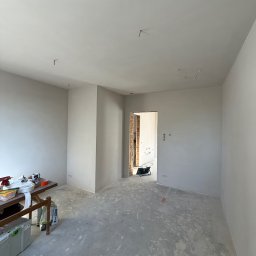 REMIGIUSZ PUKACKI REMIPOL - Pokój w trakcie prac wykończeniowych: gładkie, białe ściany, surowa podłoga, widoczne kable i narzędzia na stole. Otwór drzwiowy z odsłoniętą cegłą.
