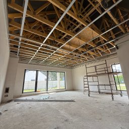 REMIGIUSZ PUKACKI REMIPOL - Montaż metalowej konstrukcji sufitu podwieszanego w surowym wnętrzu. Widoczne drewniane elementy więźby dachowej i rusztowanie. Duże okna w tle.