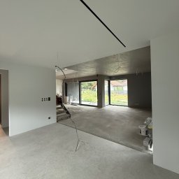 REMIGIUSZ PUKACKI REMIPOL - Surowe wnętrze domu w trakcie wykańczania: szare ściany, betonowa podłoga, widoczne puszki z farbą i kable. Minimalistyczne oświetlenie liniowe.