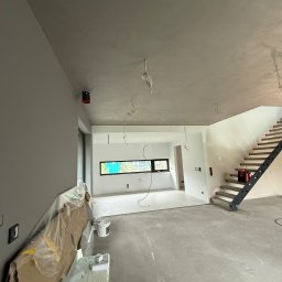 REMIGIUSZ PUKACKI REMIPOL - Surowe wnętrze w trakcie wykończenia: szpachlowane ściany, betonowa podłoga, schody z metalową konstrukcją. Widoczne kable i materiały budowlane.