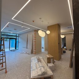 REMIGIUSZ PUKACKI REMIPOL - Wnętrze w trakcie wykańczania: widoczne płyty gipsowe, drabina, oświetlenie sufitowe LED i podwieszane lampy. Posadzka z lastryko. Prace wykończeniowe w toku.