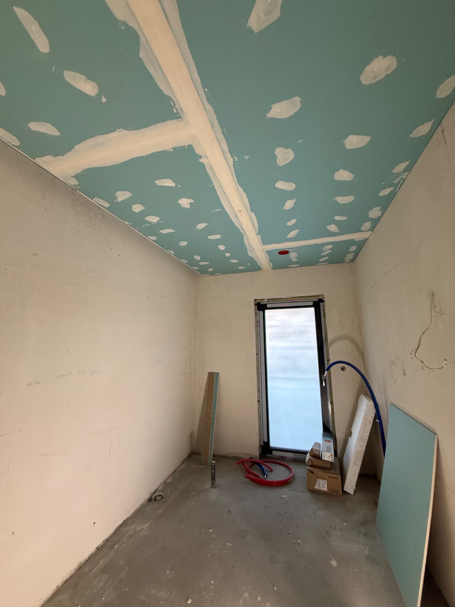 Surowy pokój w trakcie prac: płyta gipsowa na suficie z widocznymi łączeniami, ściany przygotowane do dalszych prac, materiały budowlane przy oknie.