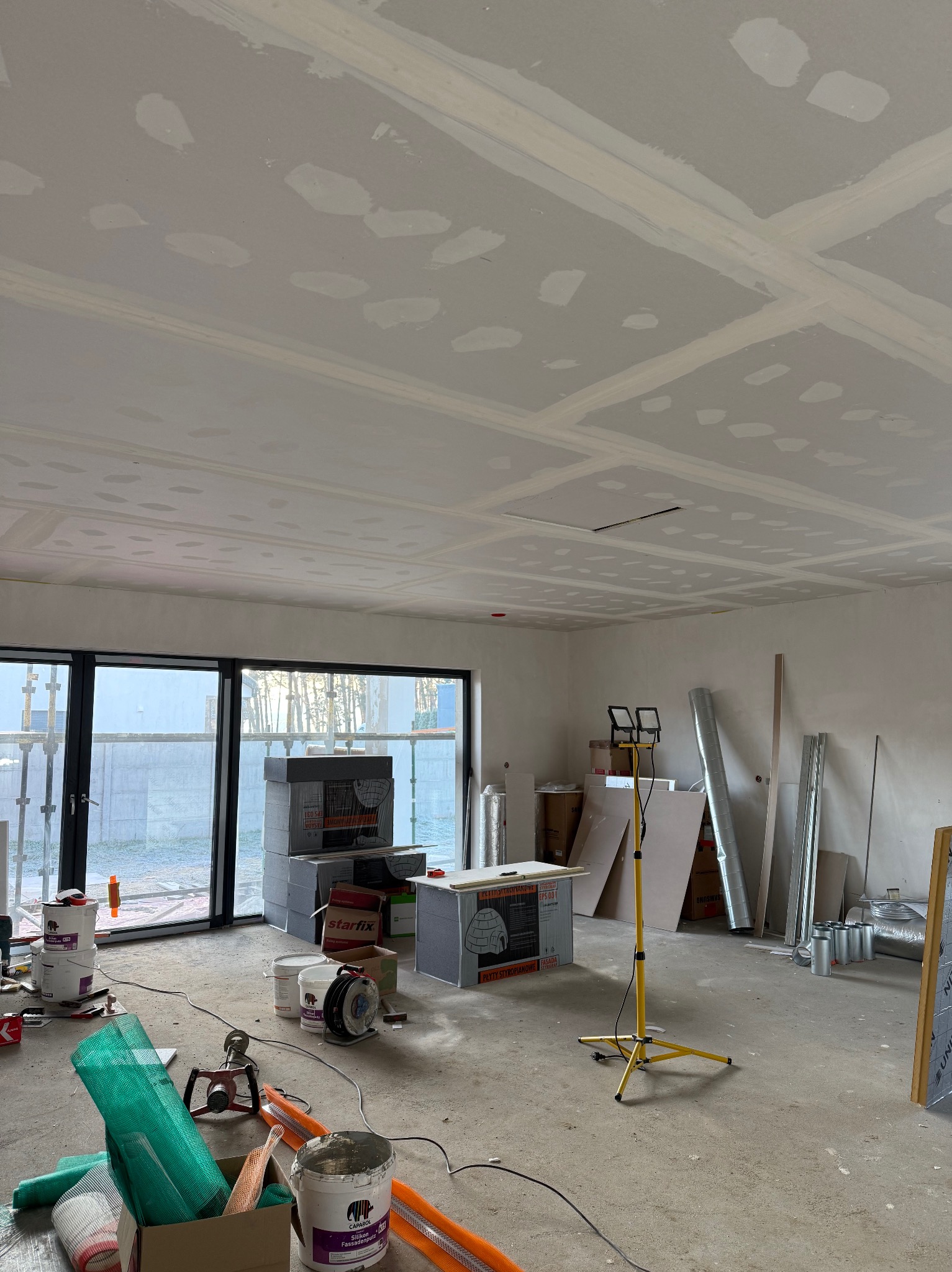 Wnętrze w trakcie prac wykończeniowych: sufit z płyt kartonowo-gipsowych po szpachlowaniu, widoczne narzędzia i materiały budowlane, lampa oświetleniowa na trójnogu.