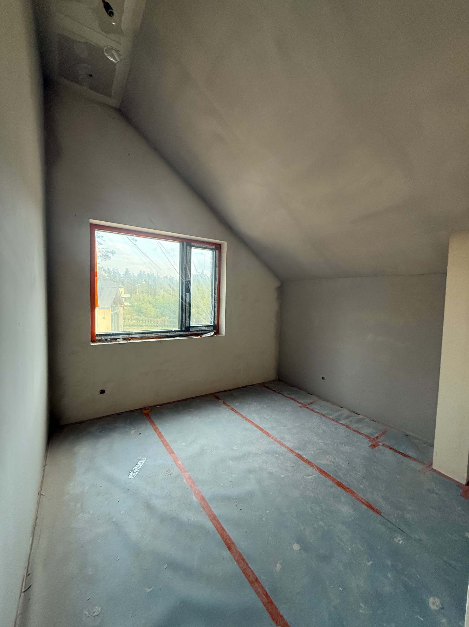 Pokój na poddaszu po szpachlowaniu ścian i sufitu, z oknem zabezpieczonym folią. Na podłodze widoczne czerwone linie wyznaczające obszar roboczy.