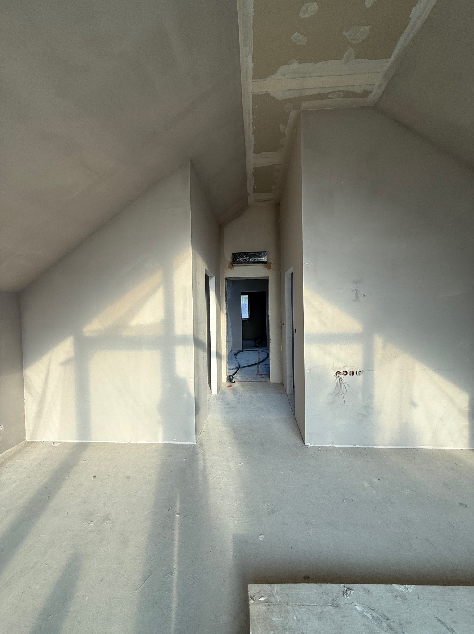 Pokój na poddaszu po gładziowaniu ścian i sufitu, widoczne przygotowane otwory na gniazdka elektryczne i fragment płyty gipsowej na suficie. Jasne, naturalne światło.