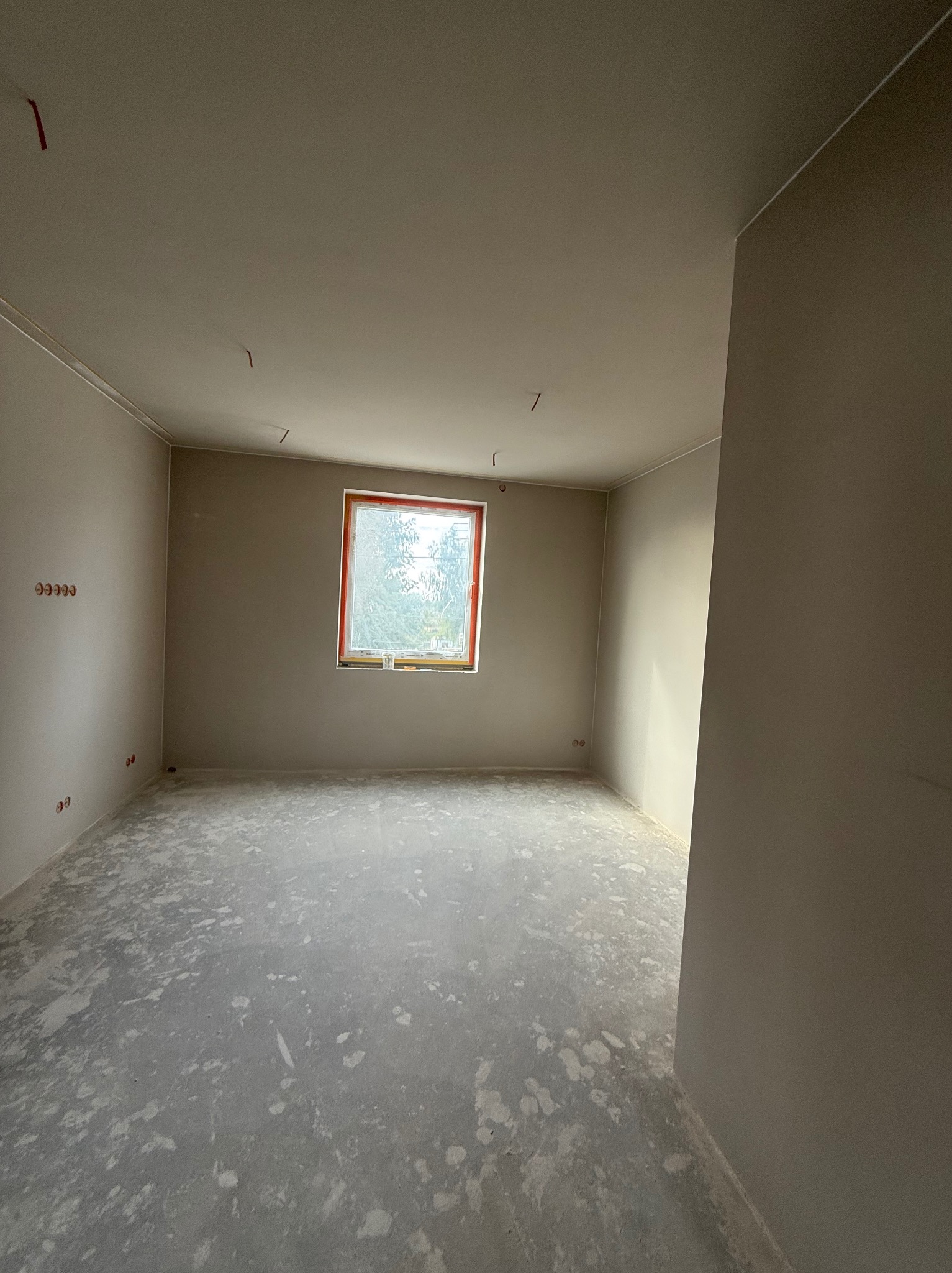 Wnętrze pokoju po gładziowaniu ścian i wylewce podłogi, z widocznymi puszkami elektrycznymi i oknem z widokiem na zieleń. Ściany w jasnym, neutralnym odcieniu.