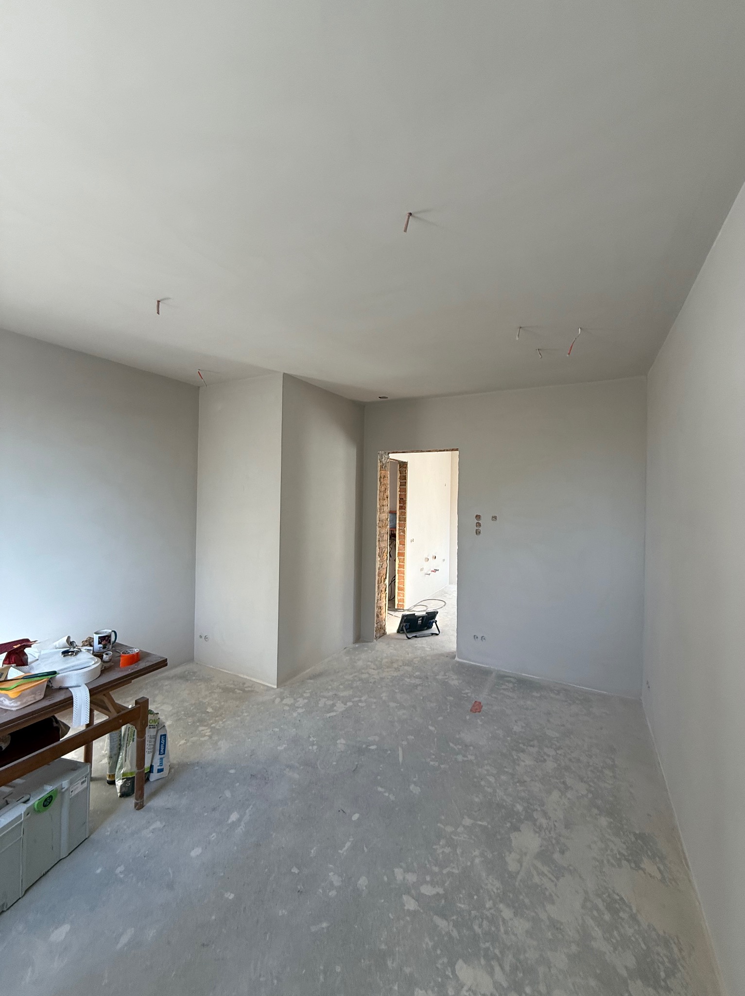 Pokój w trakcie prac wykończeniowych: gładkie, białe ściany, surowa podłoga, widoczne kable i narzędzia na stole. Otwór drzwiowy z odsłoniętą cegłą.