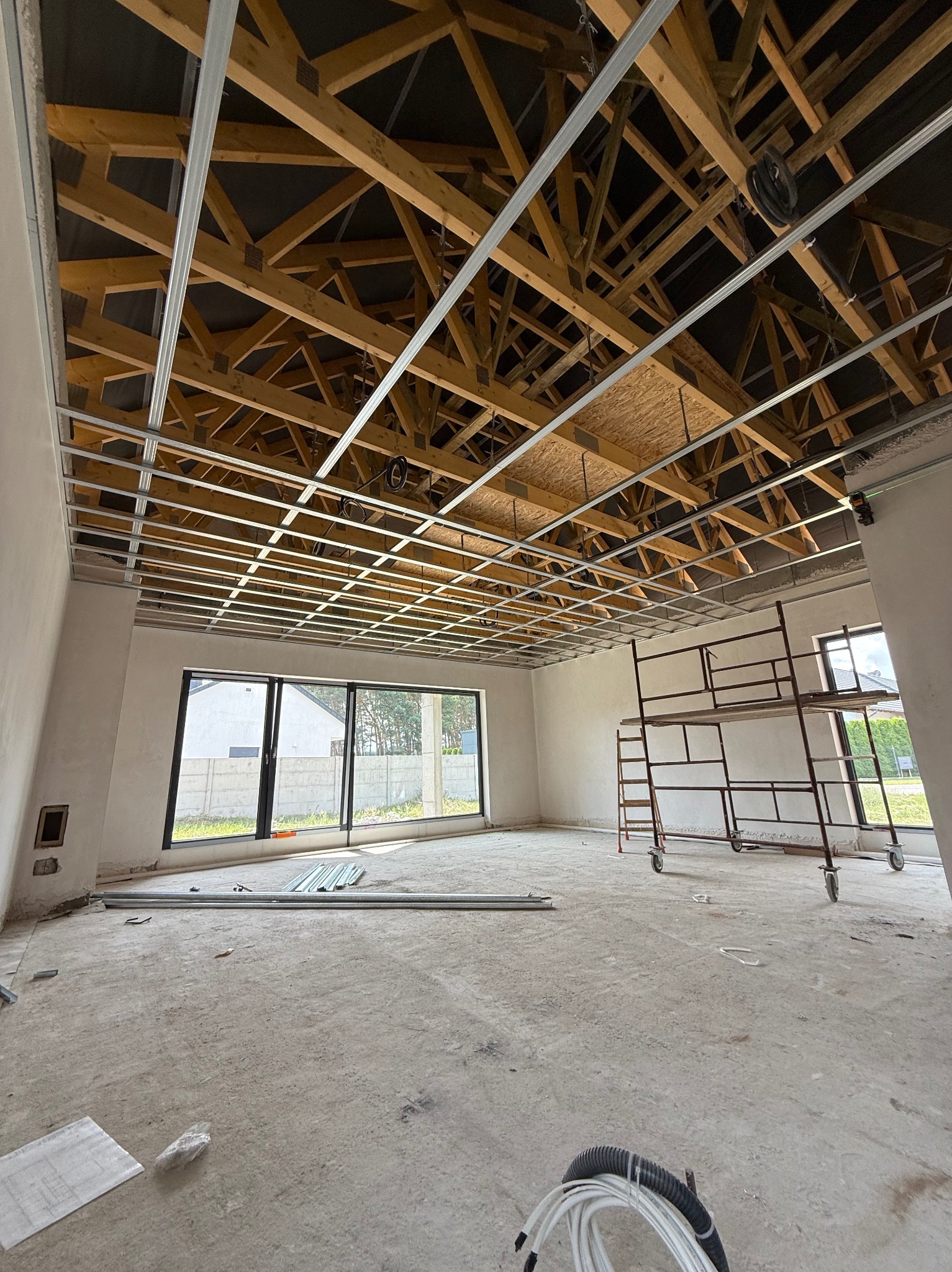 Montaż metalowej konstrukcji sufitu podwieszanego w surowym wnętrzu. Widoczne drewniane elementy więźby dachowej i rusztowanie. Duże okna w tle.