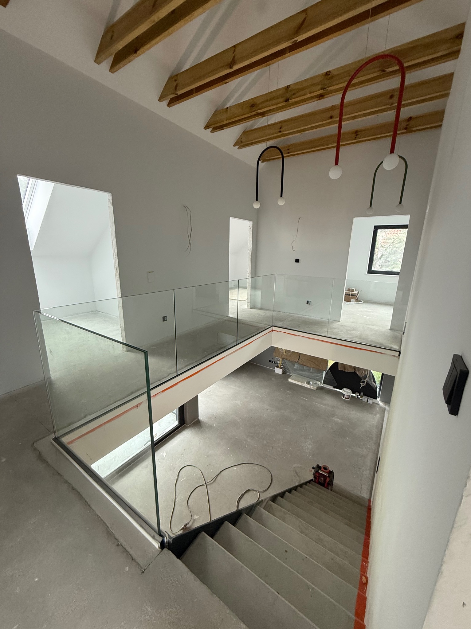 Wnętrze domu w trakcie wykańczania: surowe ściany, drewniany strop, szklane balustrady, schody i designerskie lampy. Widok z góry na dwupoziomowe wnętrze.