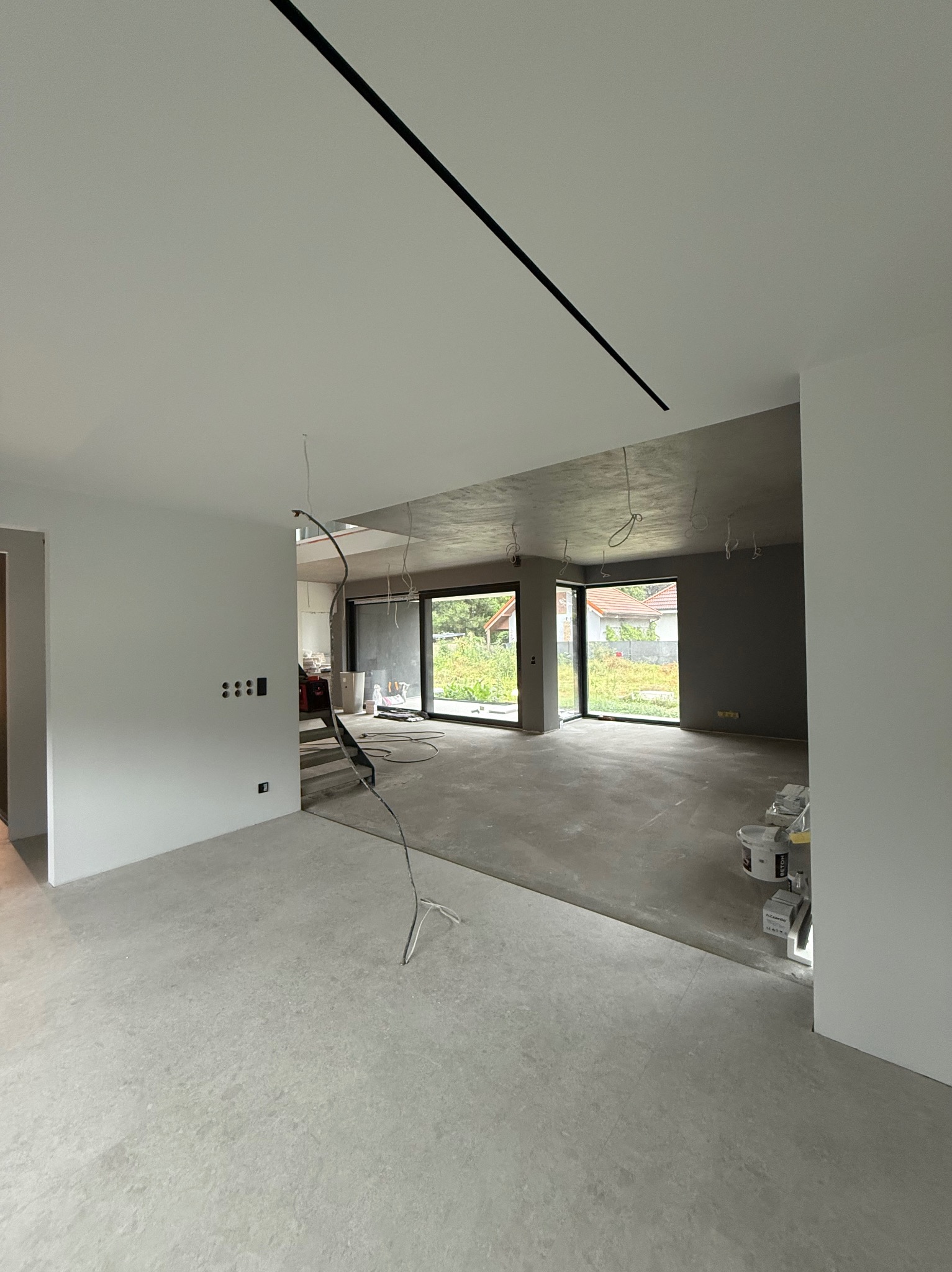 Surowe wnętrze domu w trakcie wykańczania: szare ściany, betonowa podłoga, widoczne puszki z farbą i kable. Minimalistyczne oświetlenie liniowe.
