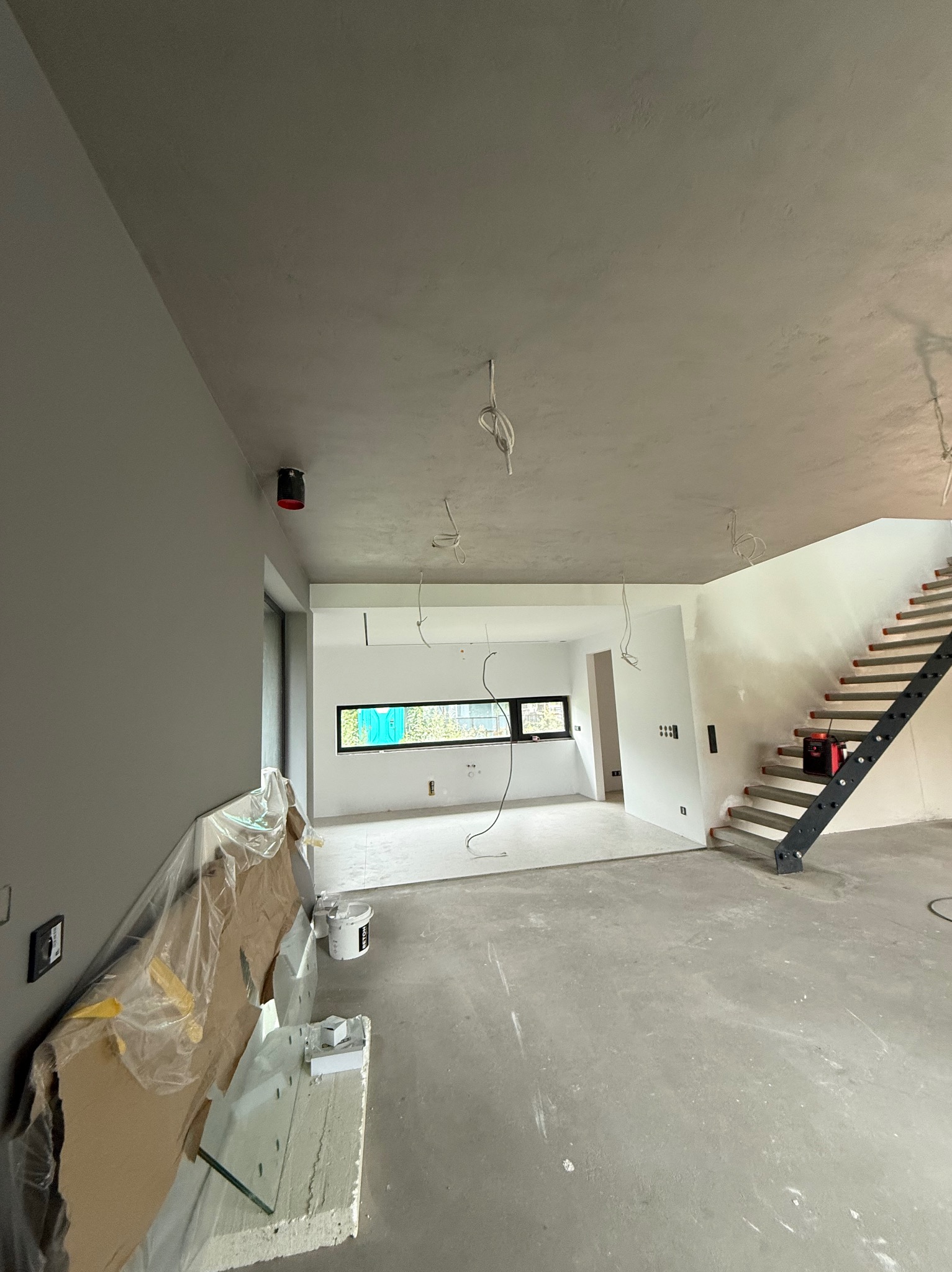 Surowe wnętrze w trakcie wykończenia: szpachlowane ściany, betonowa podłoga, schody z metalową konstrukcją. Widoczne kable i materiały budowlane.