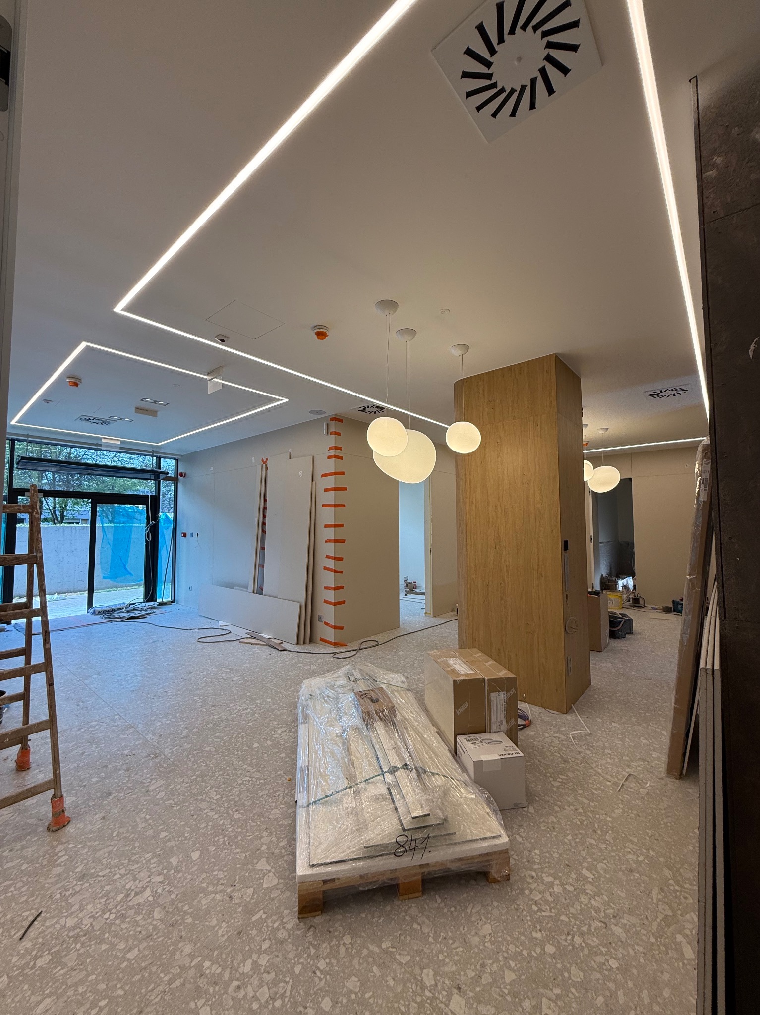 Wnętrze w trakcie wykańczania: widoczne płyty gipsowe, drabina, oświetlenie sufitowe LED i podwieszane lampy. Posadzka z lastryko. Prace wykończeniowe w toku.