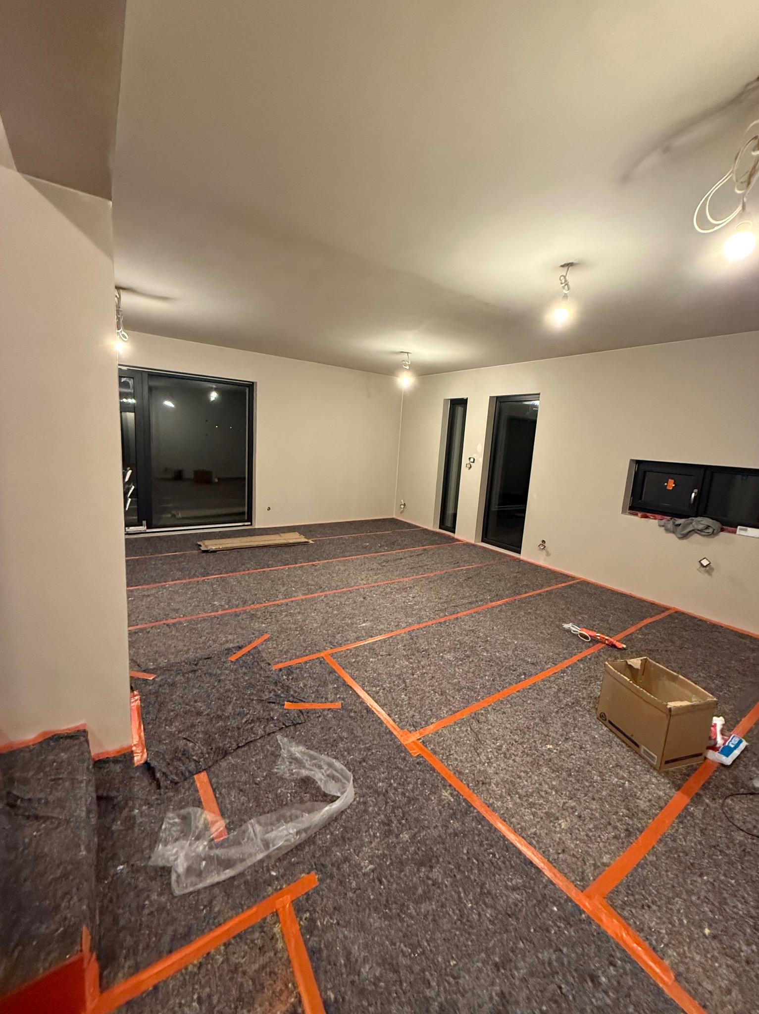 Przygotowanie do prac wykończeniowych: zabezpieczona podłoga folią i taśmą w przestronnym, jasnym wnętrzu z oknami. Widoczne narzędzia i materiały budowlane.