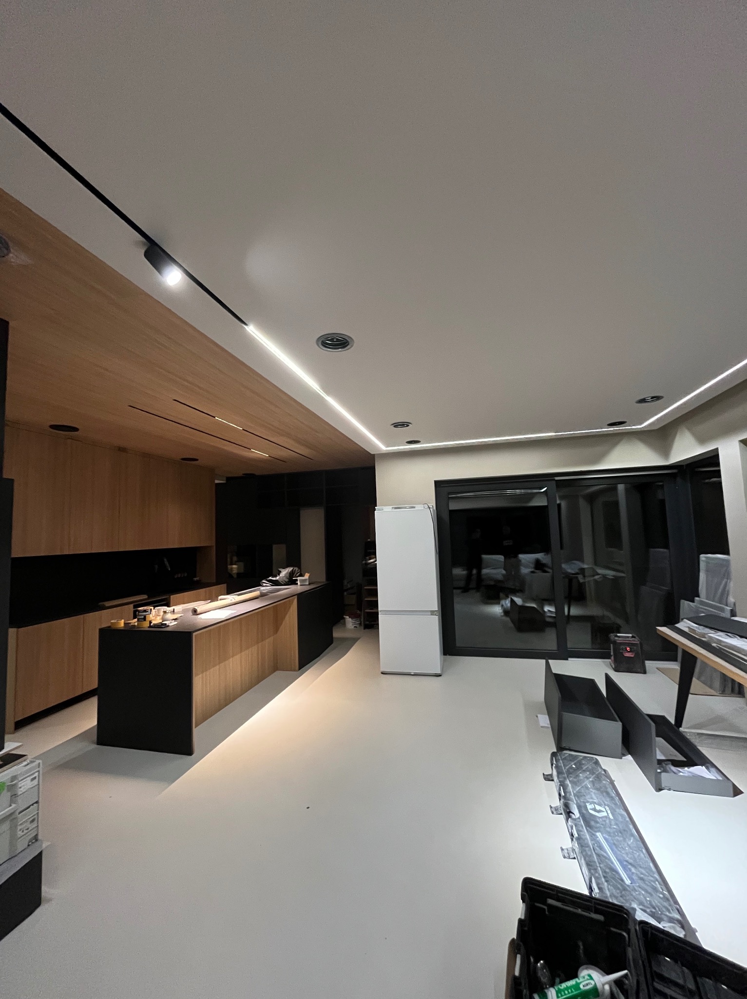 Nowoczesna kuchnia z drewnianymi szafkami i czarnym blatem, oświetlona listwami LED. Widoczne narzędzia sugerują prace wykończeniowe.