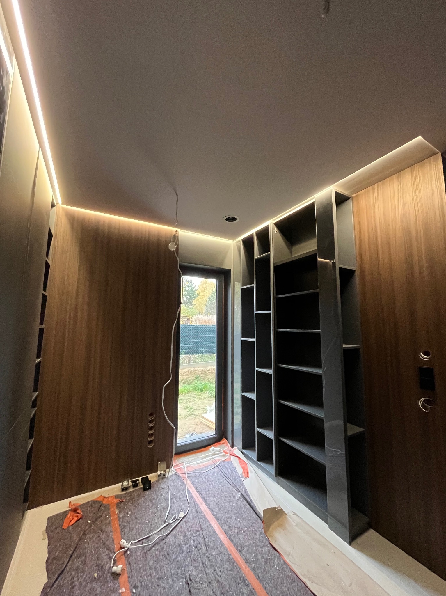 Montaż zabudowy wnękowej z półkami, widoczne oświetlenie LED, zabezpieczona podłoga. W tle okno z widokiem na zewnątrz. Prace wykończeniowe w nowoczesnym stylu.