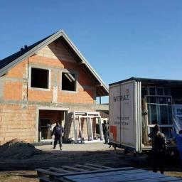 EuroMat Okna Drzwi - Dostawa okien na plac budowy domu jednorodzinnego w stanie surowym otwartym, pracownicy rozładowują z ciężarówki białe ramy okienne.