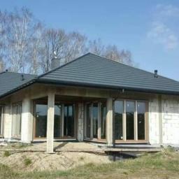 EuroMat Okna Drzwi - Surowy stan domu jednorodzinnego z szarym dachem, z widocznymi oknami PCV i drzwiami przesuwnymi, w trakcie budowy na tle drzew i błękitnego nieba, z zaparkowanym samochodem w tle.