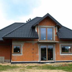 EuroMat Okna Drzwi - Surowy stan budowy domu jednorodzinnego z widocznymi oknami PCV z brązowymi ramami, drzwiami przesuwnymi i czarnym dachem z dachówki, bez elewacji zewnętrznej, z osobą pracującą w tle.