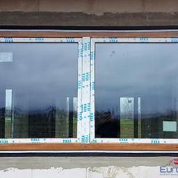 EuroMat Okna Drzwi - Zamontowane okno PCV z brązową ramą, z folią ochronną na profilach, widoczne na tle surowej ściany budynku w trakcie budowy.