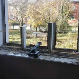 Demontaż starego okna: widok z wnętrza na częściowo usuniętą ramę okienną, z resztkami gruzu na parapecie i narzędziem akumulatorowym leżącym obok, z widokiem na jesienne drzewa i budynek na zewnątrz.