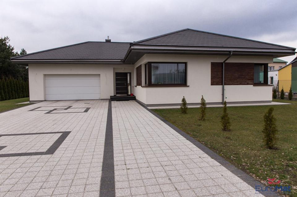 Nowoczesny dom jednorodzinny z białymi ścianami, ciemną stolarką okienną i grafitowym dachem, widziany z perspektywy podjazdu wyłożonego jasnoszarą kostką brukową z elementami grafitowymi.