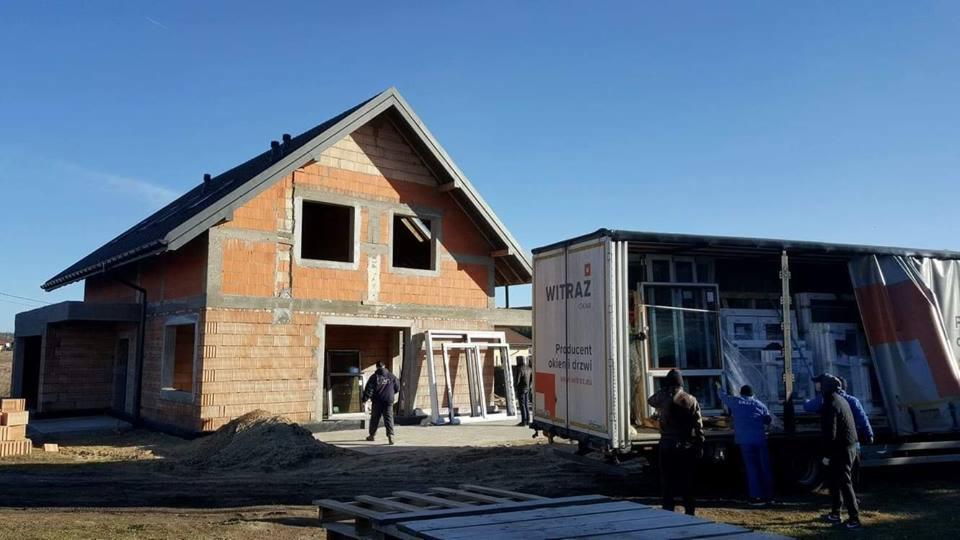 Dostawa okien na plac budowy domu jednorodzinnego w stanie surowym otwartym, pracownicy rozładowują z ciężarówki białe ramy okienne.