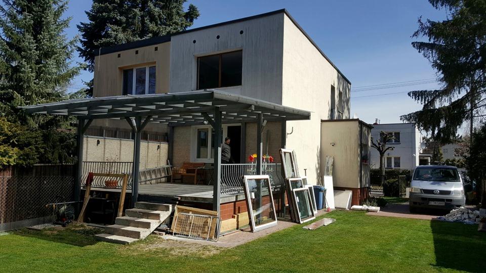 Dom jednorodzinny podczas wymiany okien, widoczne okna przygotowane do montażu na trawniku, częściowo zdemontowane stare okna, pracownik na tarasie.