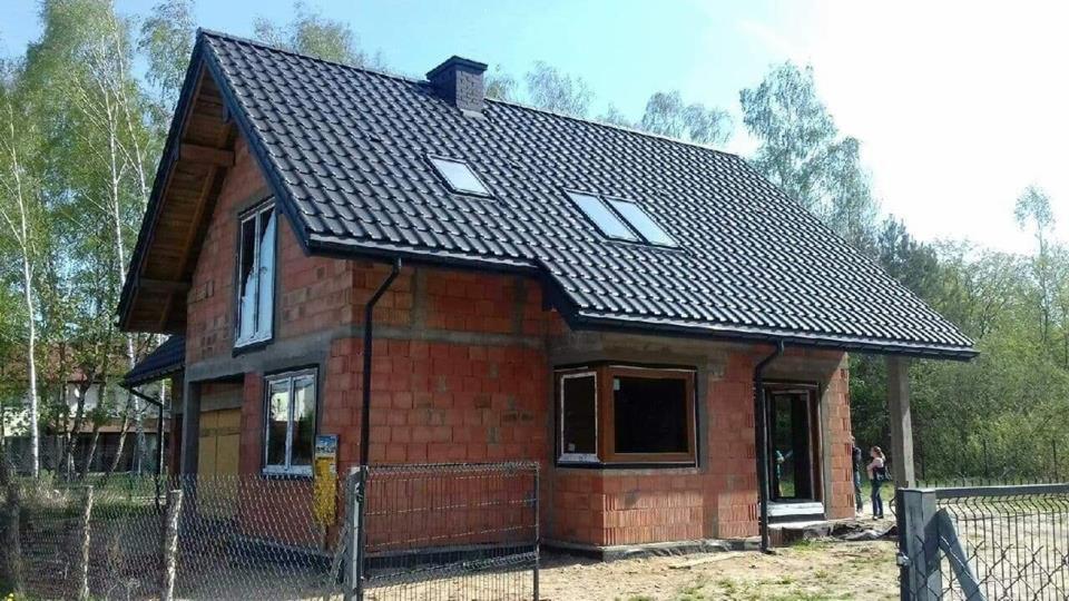 Surowy stan budynku jednorodzinnego z dachem pokrytym czarną dachówką, widoczne okna w trakcie montażu i niezabezpieczone otwory okienne, ceglany mur bez ocieplenia, ogrodzenie z siatki metalowej.