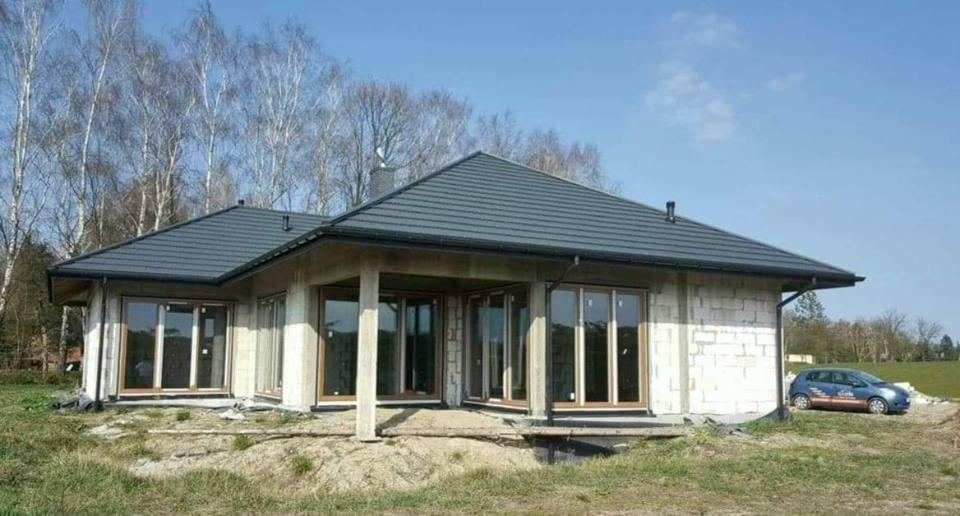 Surowy stan domu jednorodzinnego z szarym dachem, z widocznymi oknami PCV i drzwiami przesuwnymi, w trakcie budowy na tle drzew i błękitnego nieba, z zaparkowanym samochodem w tle.