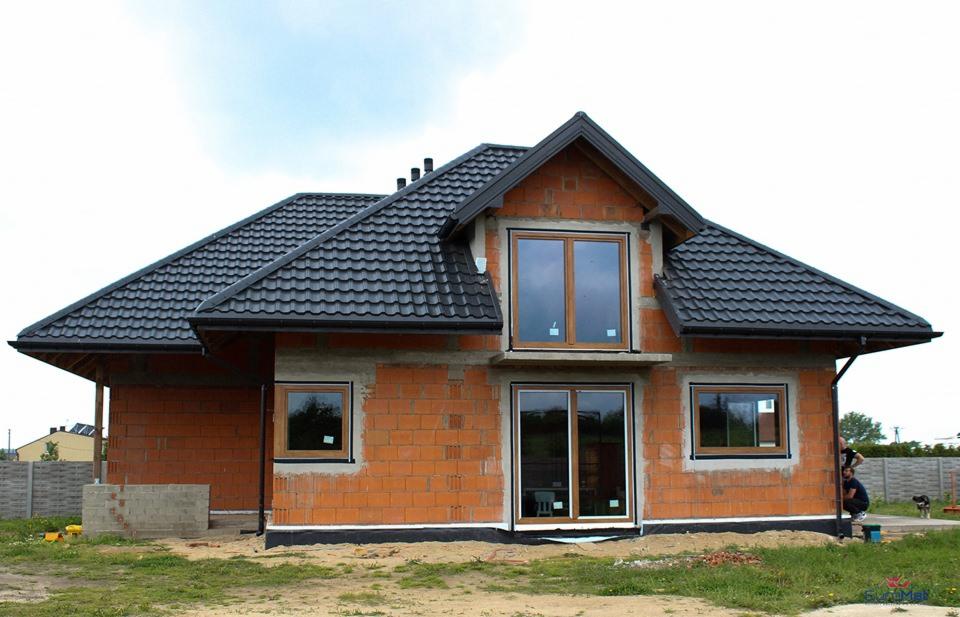 Surowy stan budowy domu jednorodzinnego z widocznymi oknami PCV z brązowymi ramami, drzwiami przesuwnymi i czarnym dachem z dachówki, bez elewacji zewnętrznej, z osobą pracującą w tle.