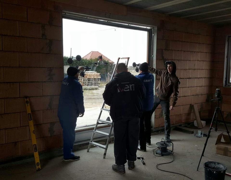 Montaż okna przez ekipę budowlaną w surowym stanie budynku, widoczne przyssawki do szkła i drabina.