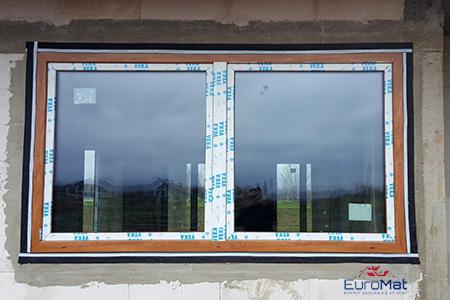 Zamontowane okno PCV z brązową ramą, z folią ochronną na profilach, widoczne na tle surowej ściany budynku w trakcie budowy.