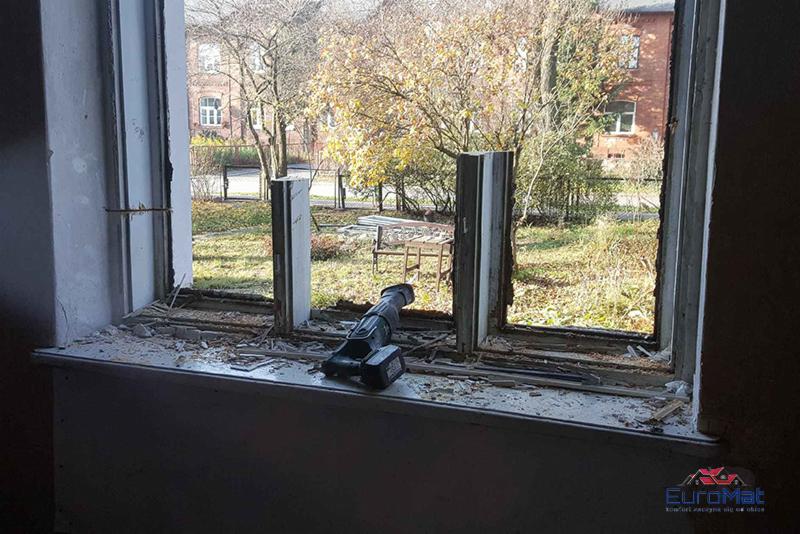Demontaż starego okna: widok z wnętrza na częściowo usuniętą ramę okienną, z resztkami gruzu na parapecie i narzędziem akumulatorowym leżącym obok, z widokiem na jesienne drzewa i budynek na zewnątrz.