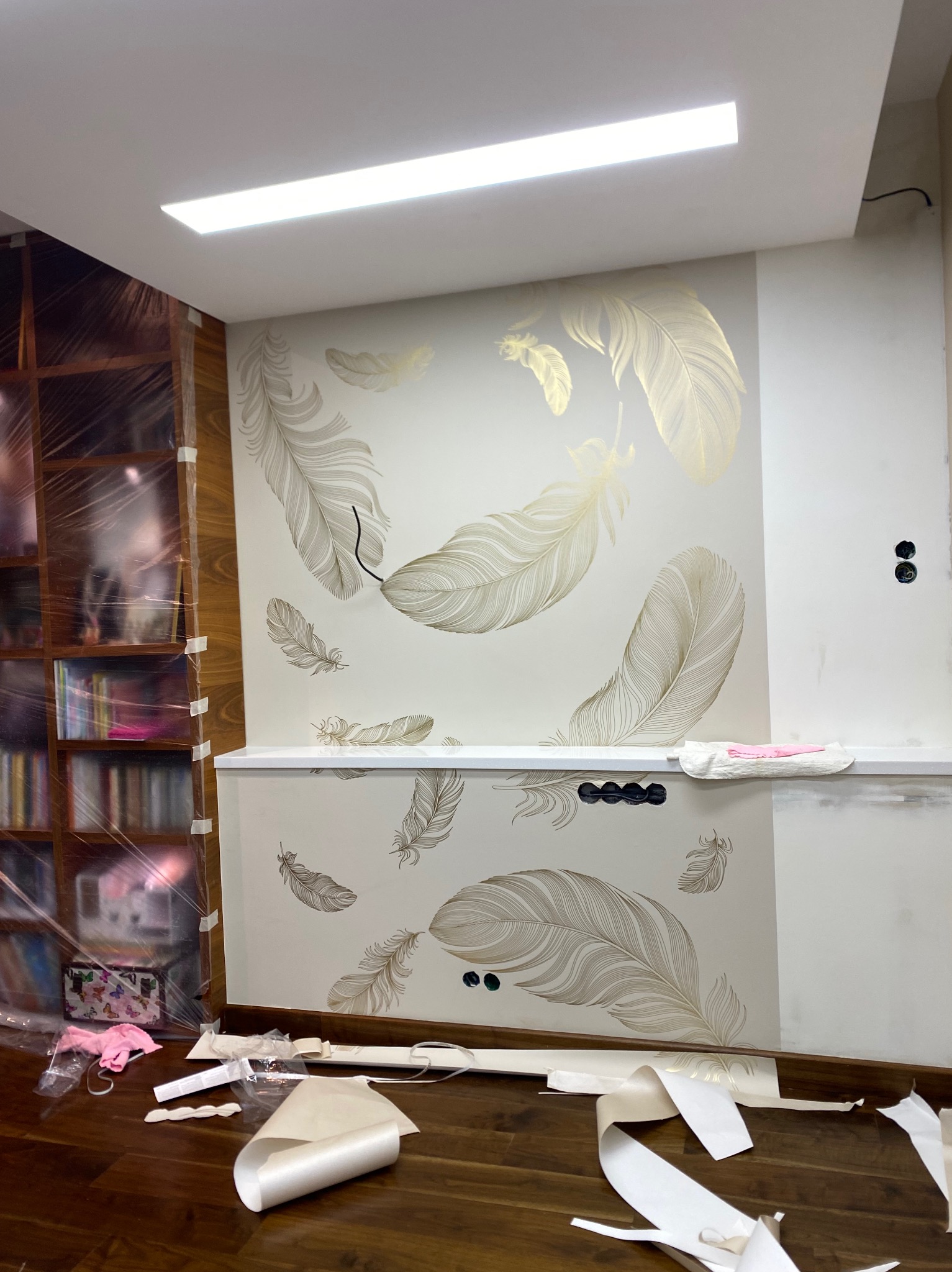 Ściana z motywem piór w odcieniach złota, częściowo pokryta tapetą. Widoczne resztki tapety na podłodze i zabezpieczony regał. Fragment gładkiej, białej ściany obok.