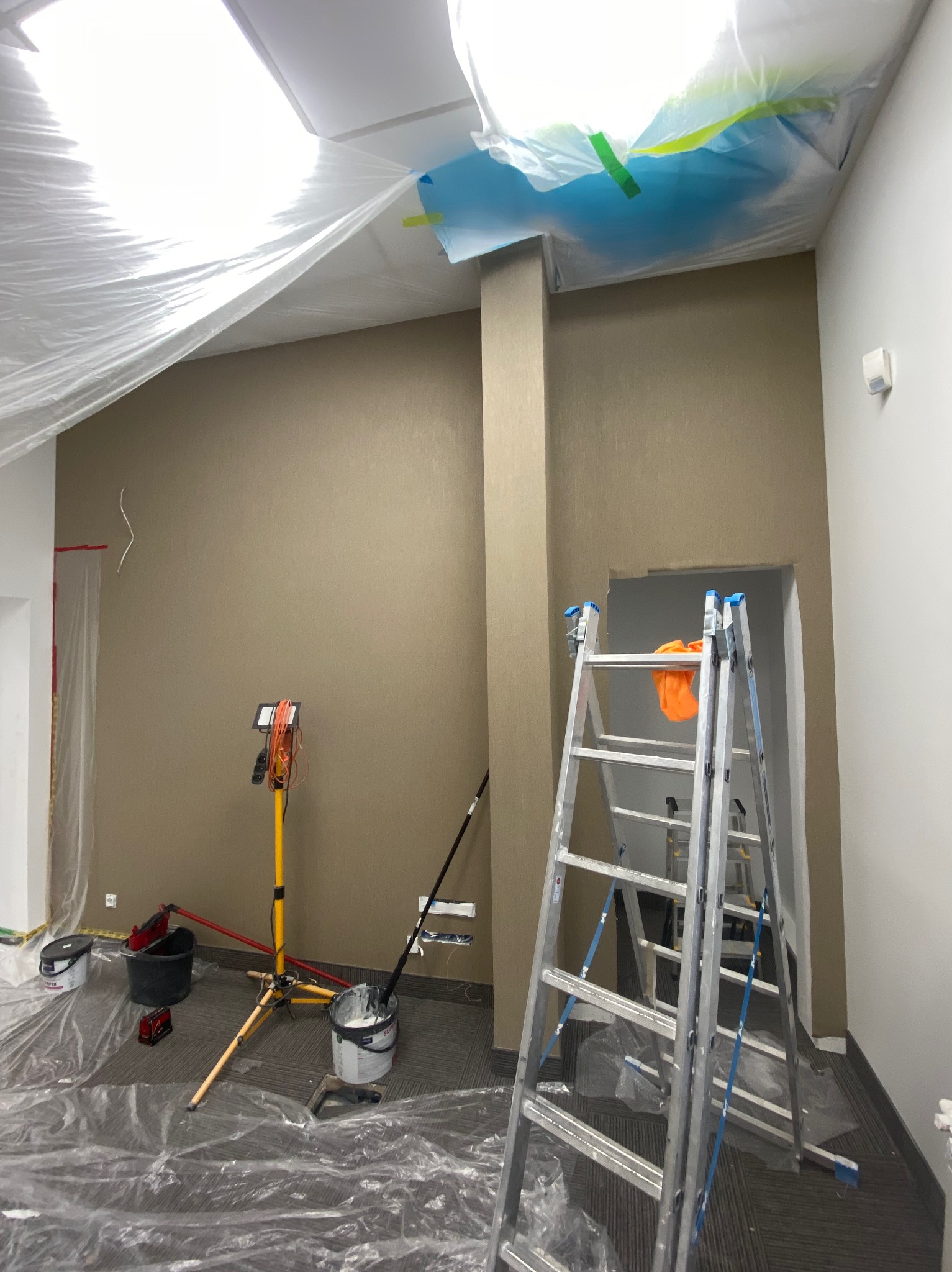 Pokój przygotowany do malowania: zabezpieczona folią sufit i podłoga, drabina, wiadro z farbą, lampa oświetleniowa. Ściana w odcieniach beżu.