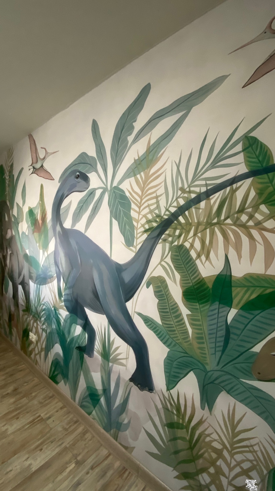 Malowidło ścienne z motywem prehistorycznym: dinozaury i roślinność tropikalna w odcieniach zieleni i brązu, oświetlone naturalnym światłem, widok z perspektywy podłogi.