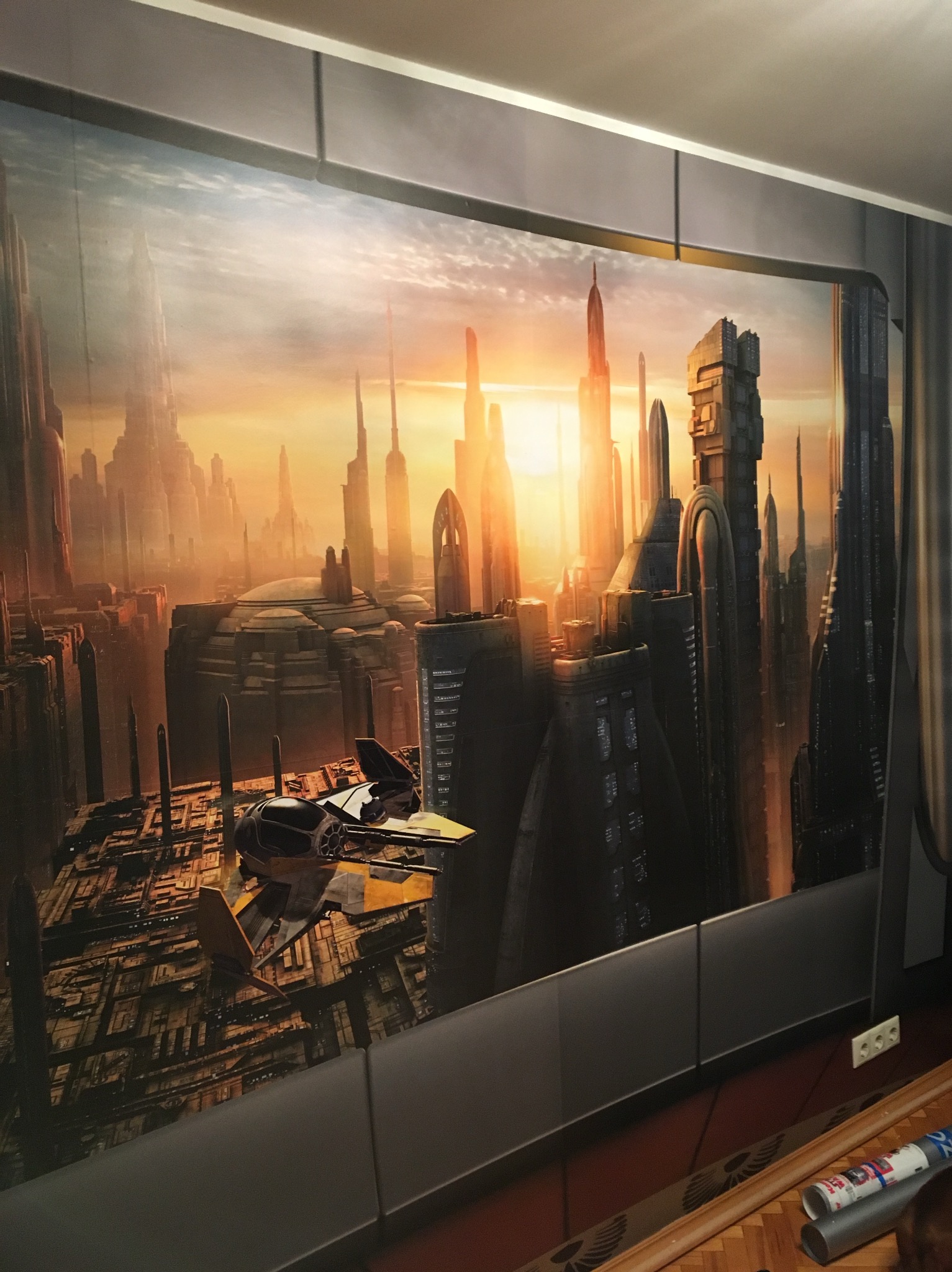 Tapeta z motywem futurystycznego miasta i statku kosmicznego, naklejona na ścianie. Widoczne łączenia tapety i fragment podłogi z akcesoriami do tapetowania.