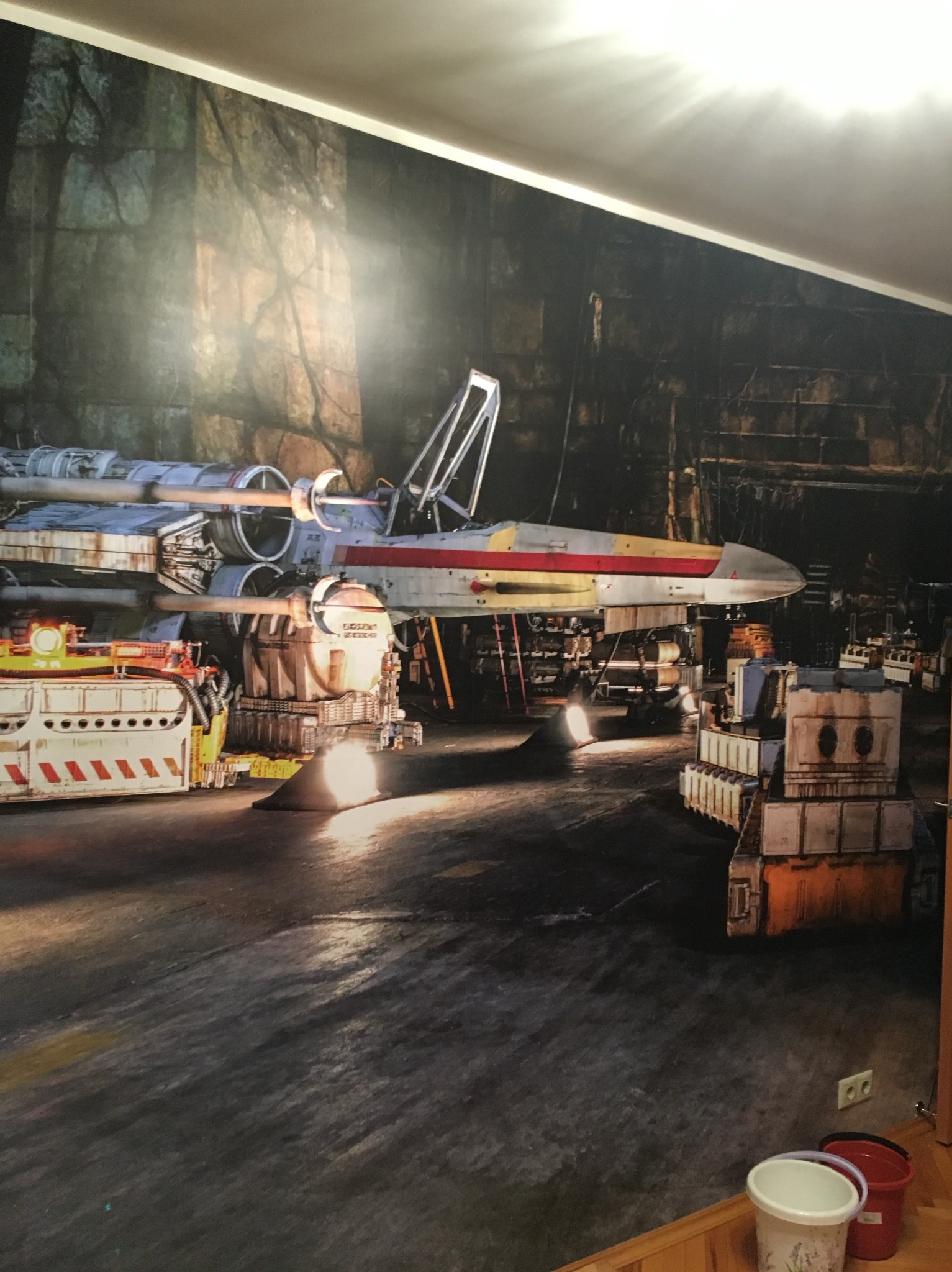 Tapeta z motywem hangaru i myśliwca X-Wing, z widocznymi detalami tekstury ściany i elementami wyposażenia. Realistyczne odwzorowanie, ciemna kolorystyka.