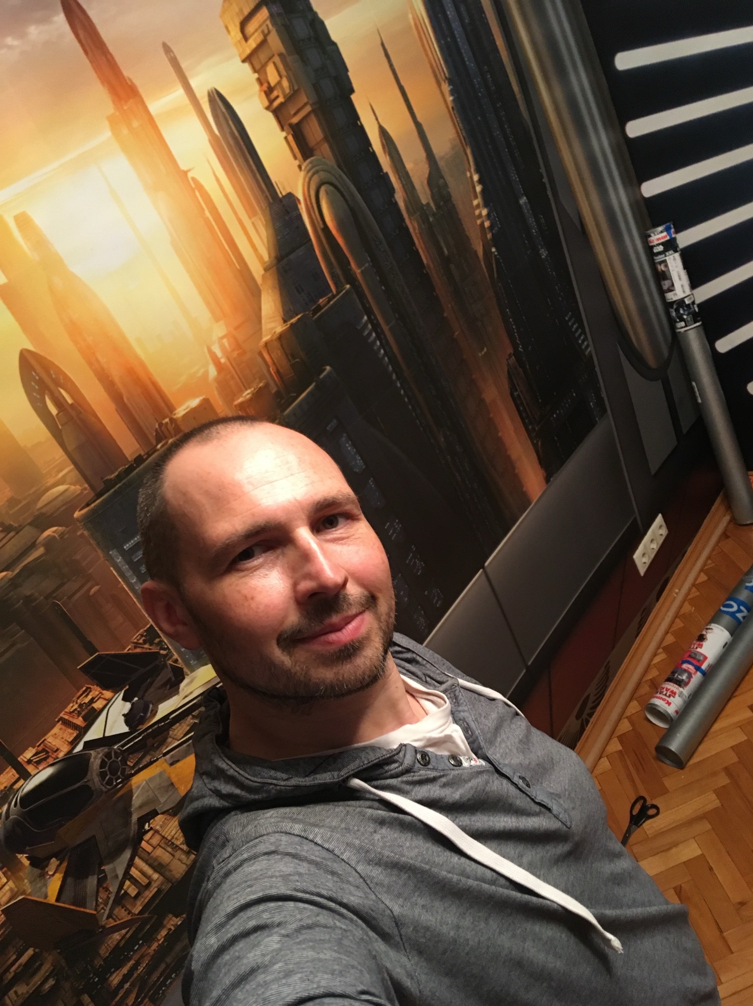 Mężczyzna na tle ściany z fototapetą przedstawiającą futurystyczne miasto. Widoczne tuby po tapecie i nożyczki na parkiecie. Ujęcie z dołu, lekko z boku.