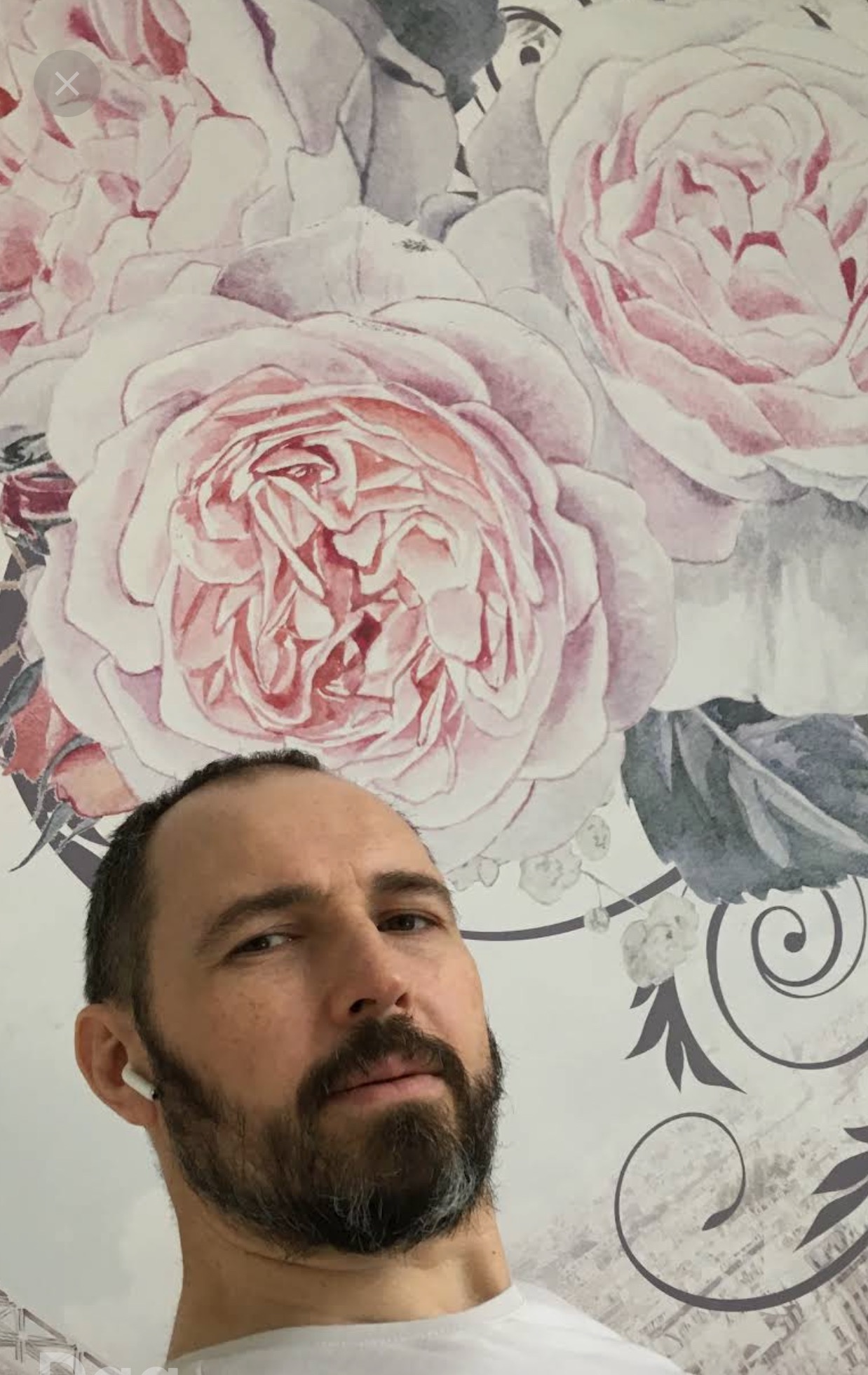 Mężczyzna z brodą na tle ściany z tapetą w duże, różowe róże. Widoczny fragment twarzy i ramienia. Tapeta w stylu akwareli z szarymi elementami.