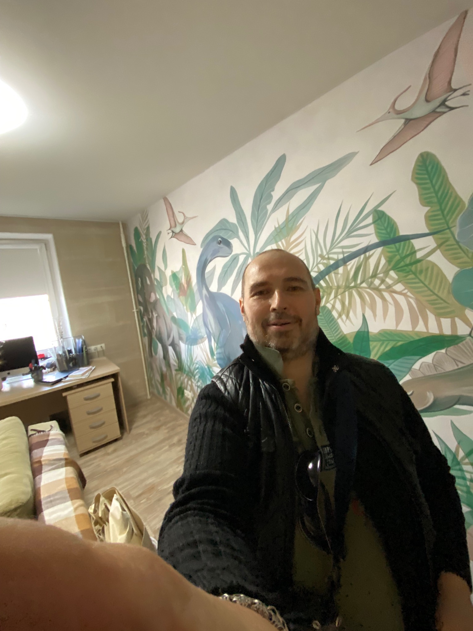 Mężczyzna na tle ściany z motywem dżungli i dinozaurów, prawdopodobnie świeżo po tapetowaniu. Widoczny fragment pokoju z biurkiem i łóżkiem.