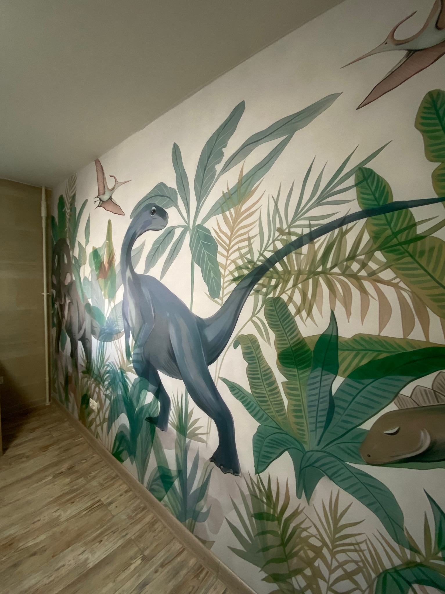 Malowidło ścienne w pokoju dziecięcym: motyw dżungli z dinozaurami w odcieniach zieleni i szarości. Widok z perspektywy podłogi, ciepłe oświetlenie.