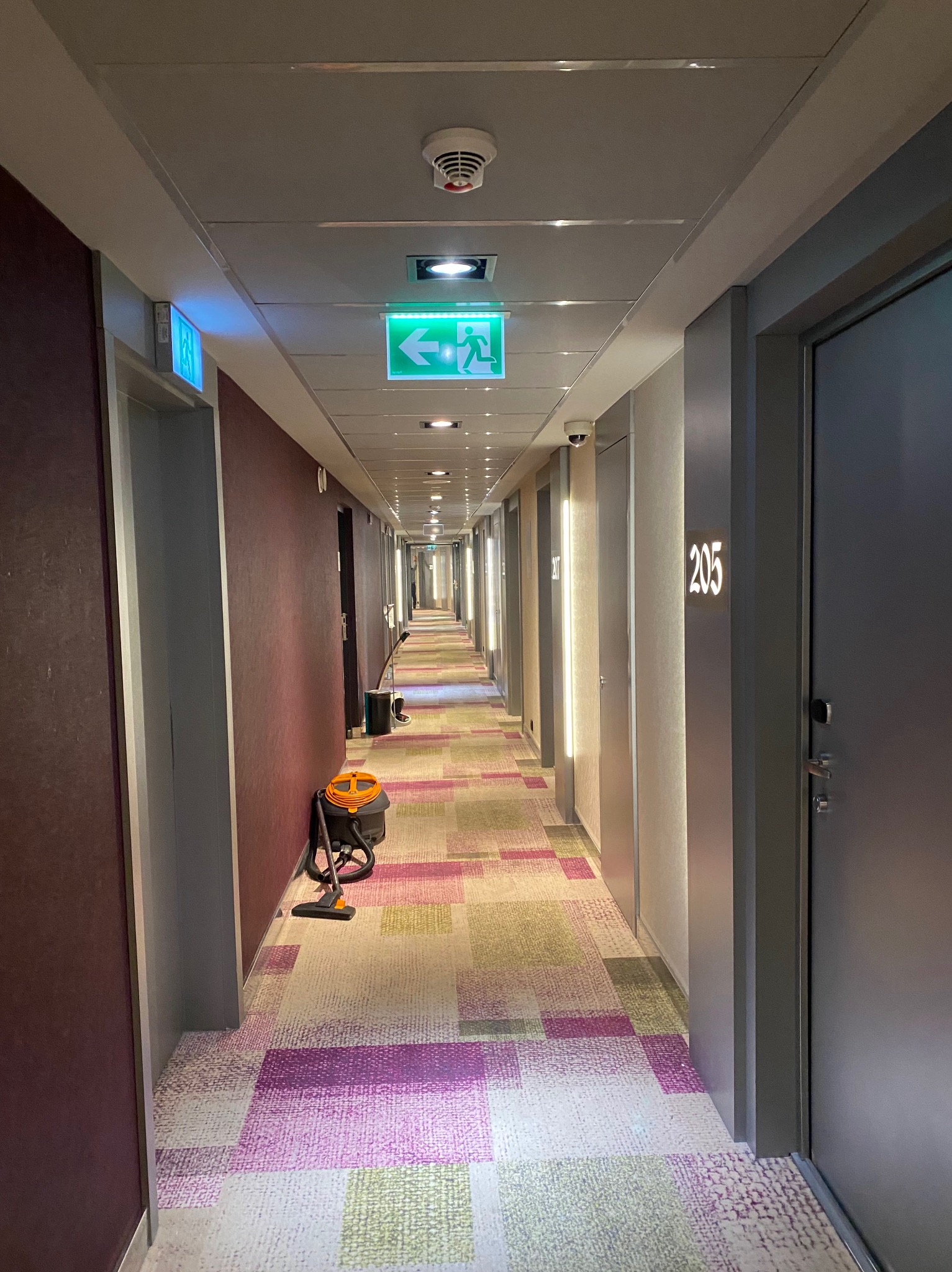 Długi korytarz hotelowy z wzorzystą wykładziną, oświetleniem LED i numerami pokoi. W tle widoczny odkurzacz, sugerujący prace porządkowe.